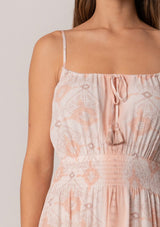 [Color: Natural/Peach] A close up front facing image of a brunette model wearing a summer mini dress in a pink bohemian print. With adjustable spaghetti straps, a scoop neckline, a tiered mini skirt, a smocked elastic waist, and a drawstring front detail with tassel ties. 