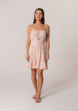 [Color: Natural/Peach] A front facing image of a brunette model wearing a summer mini dress in a pink bohemian print. With adjustable spaghetti straps, a scoop neckline, a tiered mini skirt, a smocked elastic waist, and a drawstring front detail with tassel ties. 