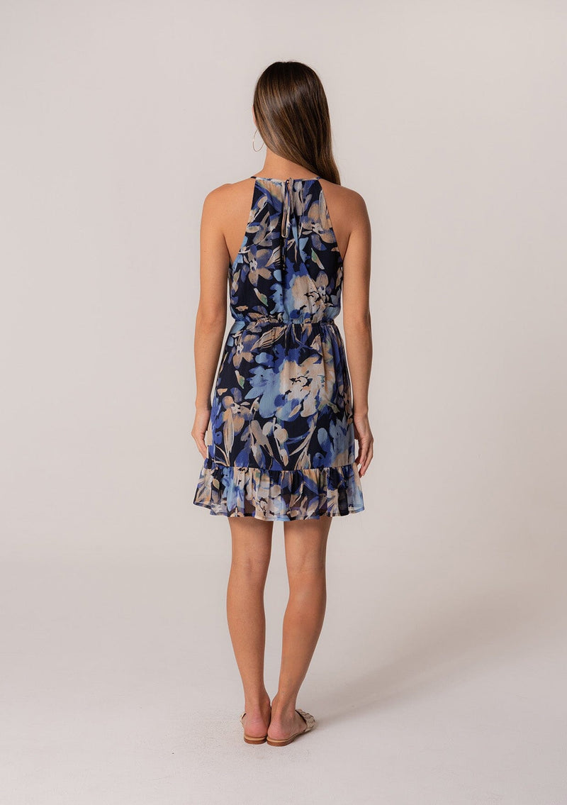 [Color: Navy/Light Blue] A back facing image of a brunette model wearing a sleeveless summer mini dress in a blue floral print. With a surplice v neckline, a tiered faux wrap skirt, an elastic waist, and a back keyhole with tie closure. 