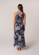 [Color: Navy/Light Blue] A back facing image of a brunette model wearing a sleeveless summer chiffon maxi dress in a blue floral print. With a v neckline in the front and back, a flowy long skirt with a side slit, an open back with tie closure, and a half smocked elastic waist. 