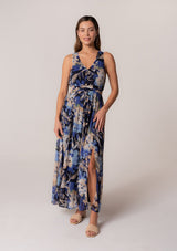 [Color: Navy/Light Blue] A front facing image of a brunette model wearing a sleeveless summer chiffon maxi dress in a blue floral print. With a v neckline in the front and back, a flowy long skirt with a side slit, an open back with tie closure, and a half smocked elastic waist. 