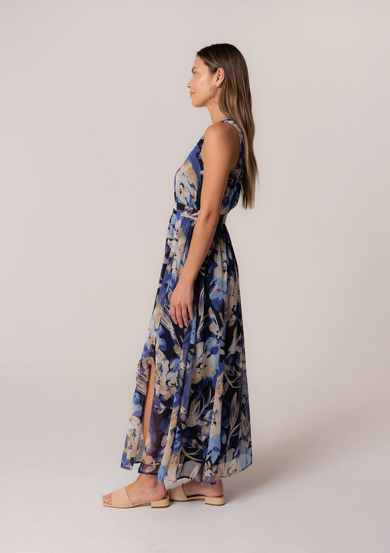 [Color: Navy/Light Blue] A side facing image of a brunette model wearing a sleeveless summer chiffon maxi dress in a blue floral print. With a v neckline in the front and back, a flowy long skirt with a side slit, an open back with tie closure, and a half smocked elastic waist. 