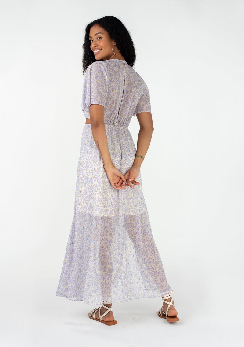 [Color: Natural/Lilac] A back facing image of a brunette model wearing a sheer chiffon bohemian maxi dress in an off white and light purple leaf print. With gold clip dot details, short flutter sleeves, a v neckline, side waist cut outs, and a back keyhole. 