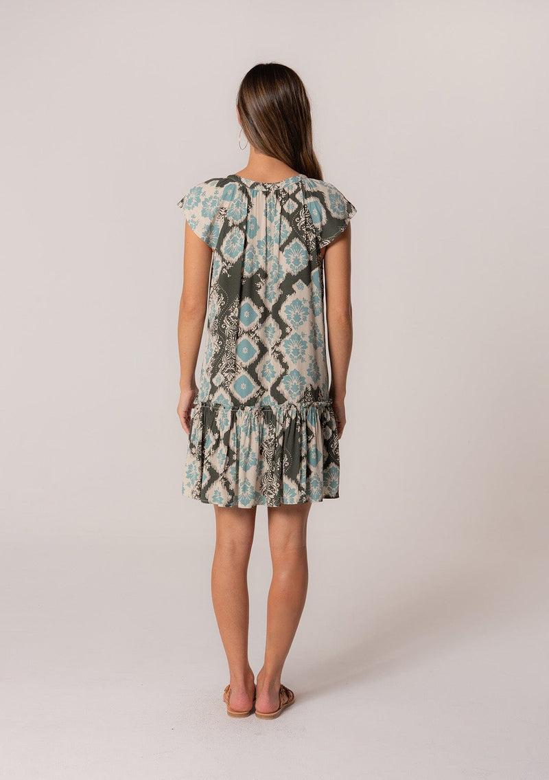 [Color: Black/Aqua] A back facing image of a brunette model wearing a lightweight summer mini dress in a green bohemian print. With short flutter sleeves, a split v-neckline with ties, and a ruffle trimmed tiered skirt. 
