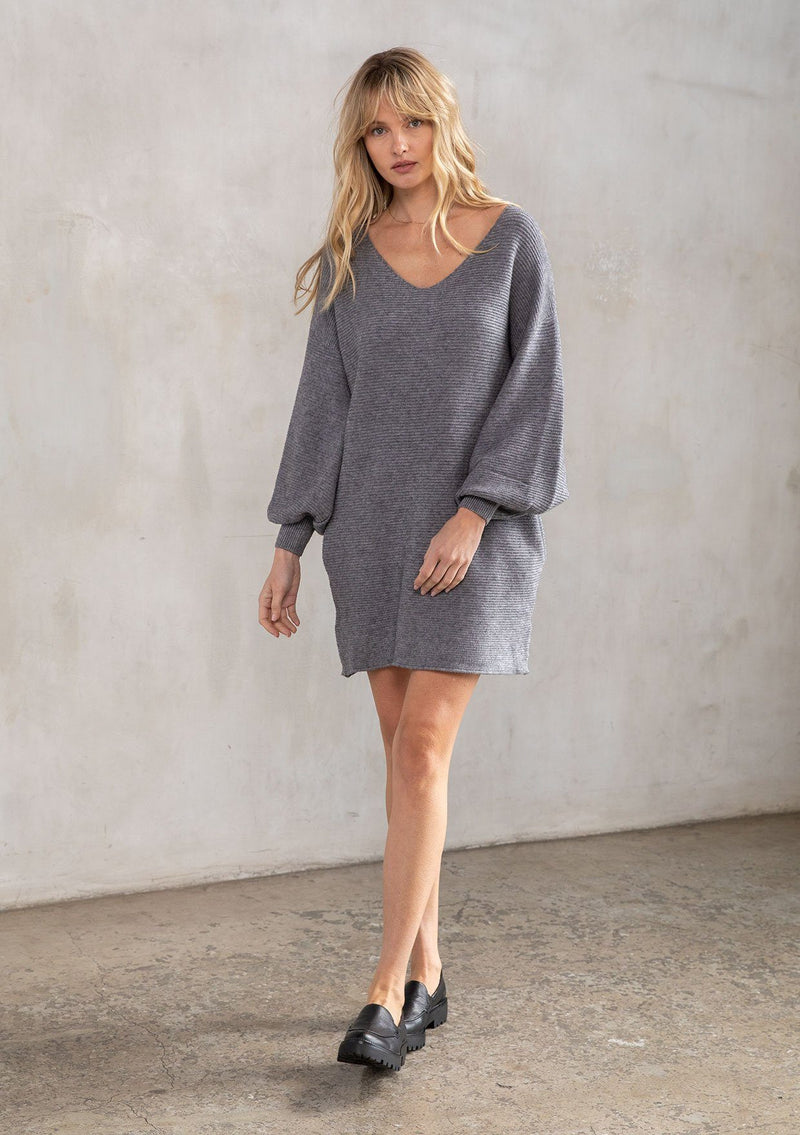 [Color: Heather Charcoal] A model wearing a heather charcoal grey ribbed knit mini sweater dress. With long bohemian balloon sleeves, side pockets, a loose relaxed fit, and a v neckline. 