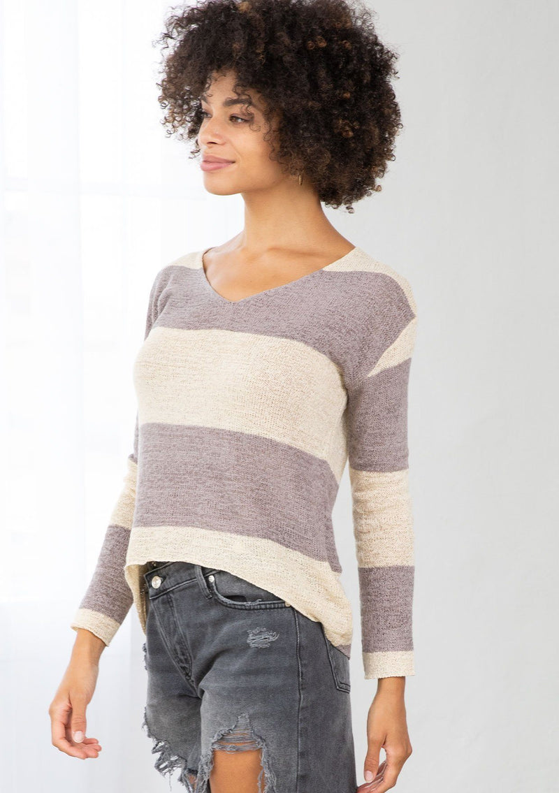 [Color: Dusty Orchid] A model wearing a dusty purple color block stripe sweater. With a wide v neckline, long sleeves, a dropped shoulder, and a high low hemline. 