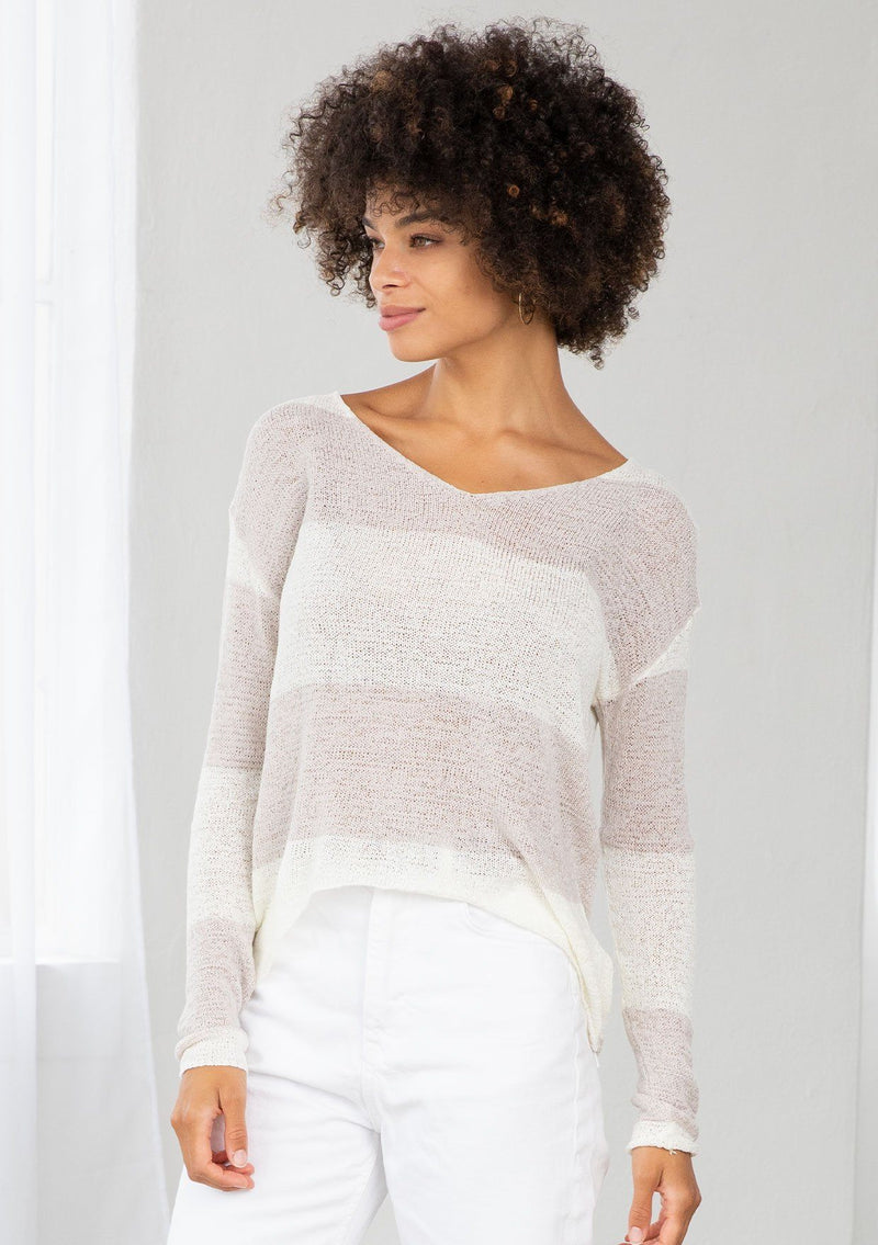 [Color: Silver/Ivory] A model wearing a silver and ivory color block stripe sweater. With a wide v neckline, long sleeves, a dropped shoulder, and a high low hemline. 