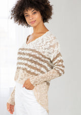 [Color: Ivory/Cement] A model wearing an ivory and sand striped color block lightweight bohemian beach sweater. With an open tie back detail, long sleeves, a dropped shoulder, and a high low hemline.