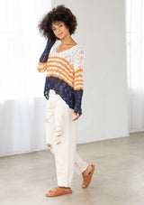 [Color: Ivory/Orange] A model wearing an ivory and orange striped color block lightweight bohemian beach sweater. With an open tie back detail, long sleeves, a dropped shoulder, and a high low hemline.