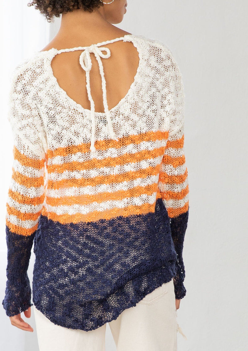 [Color: Ivory/Orange] A model wearing an ivory and orange striped color block lightweight bohemian beach sweater. With an open tie back detail, long sleeves, a dropped shoulder, and a high low hemline.
