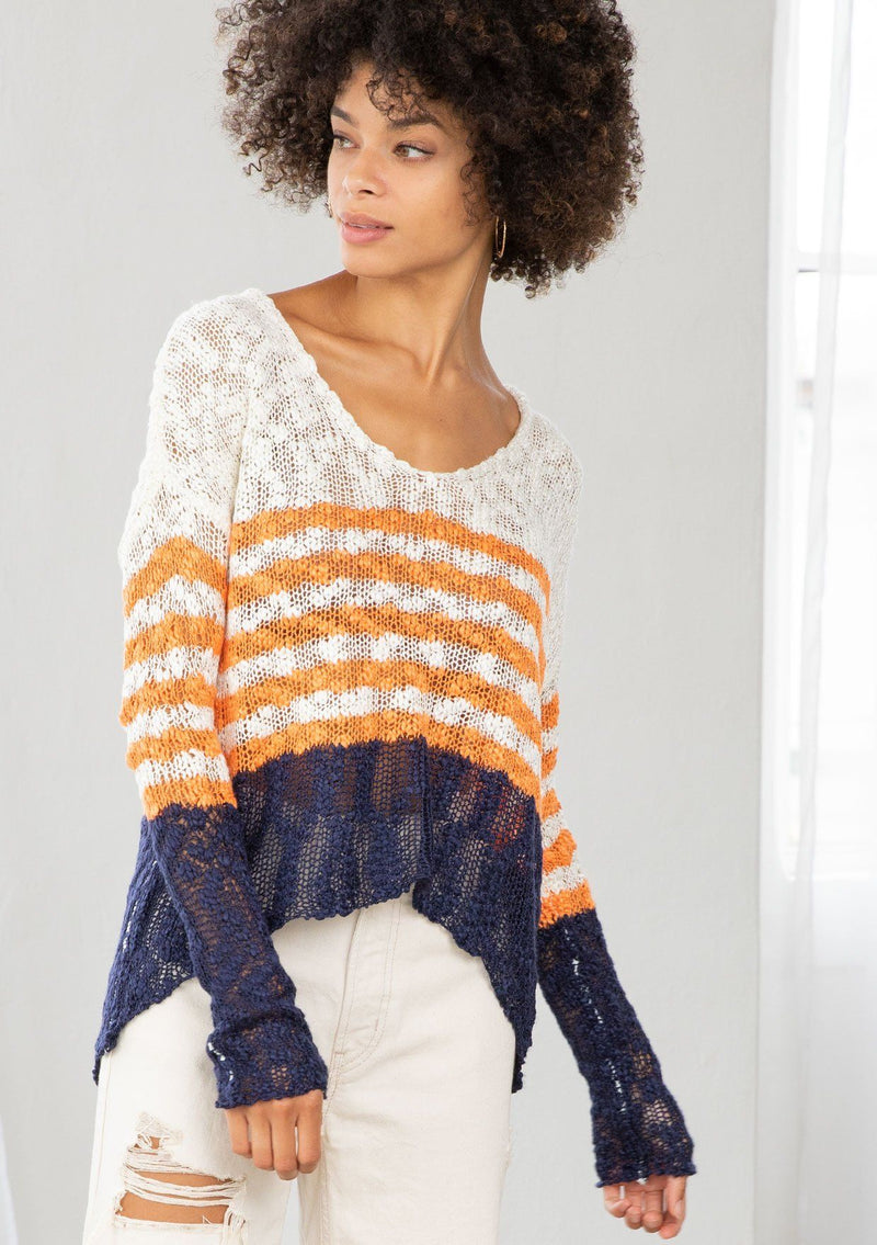 [Color: Ivory/Orange] A model wearing an ivory and orange striped color block lightweight bohemian beach sweater. With an open tie back detail, long sleeves, a dropped shoulder, and a high low hemline.