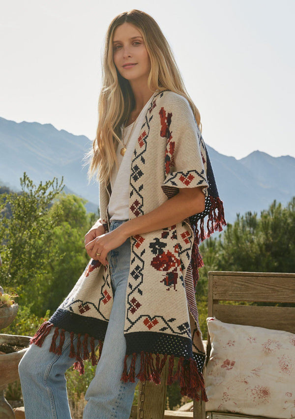 [Color: Floral] A model wearing a red and blue floral sweater poncho with fringed detail and a contrast open knit back. 