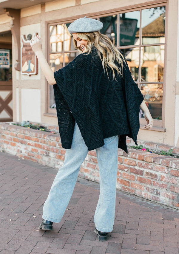 [Color: Black] A blond woman outside wearing an ultra soft chunky cable knit poncho sweater. Featuring a cozy cowl neckline.