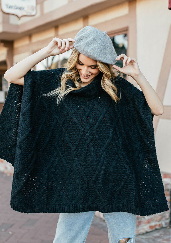 [Color: Black] A blond woman outside wearing an ultra soft chunky cable knit poncho sweater. Featuring a cozy cowl neckline.