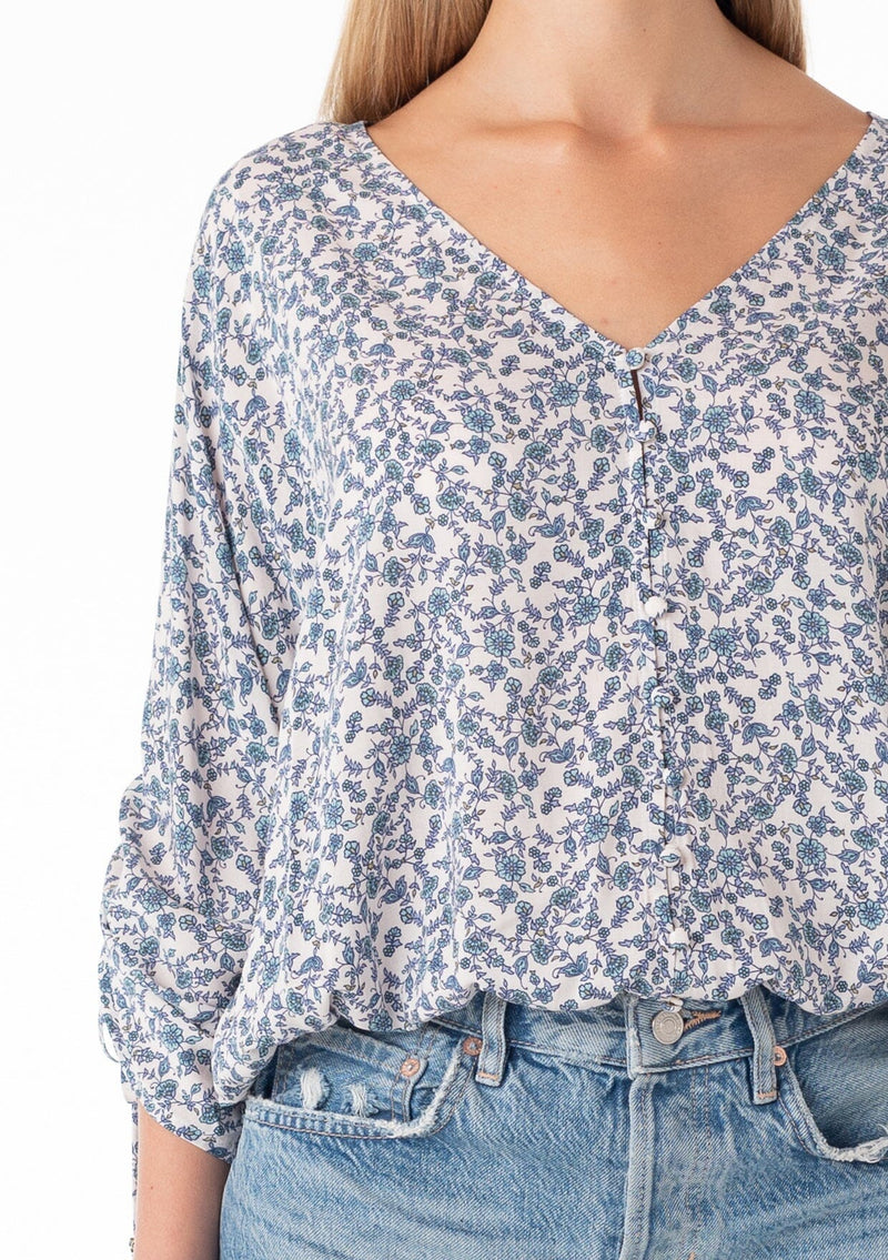[Color: Ivory/Blue] A close up front facing image of a blonde model wearing a bohemian blouse in a blue floral print. With three quarter length sleeves, a gathered drawstring sleeve detail with ties, a v neckline, a self covered button front, and a relaxed fit. 