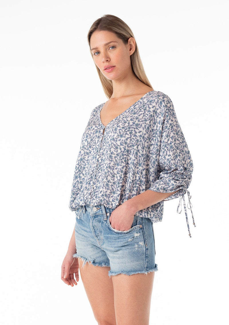 [Color: Ivory/Blue] A half body front facing image of a blonde model wearing a bohemian blouse in a blue floral print. With three quarter length sleeves, a gathered drawstring sleeve detail with ties, a v neckline, a self covered button front, and a relaxed fit. 