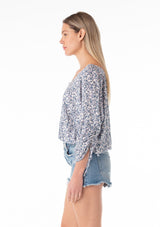 [Color: Ivory/Blue] A side facing image of a blonde model wearing a bohemian blouse in a blue floral print. With three quarter length sleeves, a gathered drawstring sleeve detail with ties, a v neckline, a self covered button front, and a relaxed fit. 