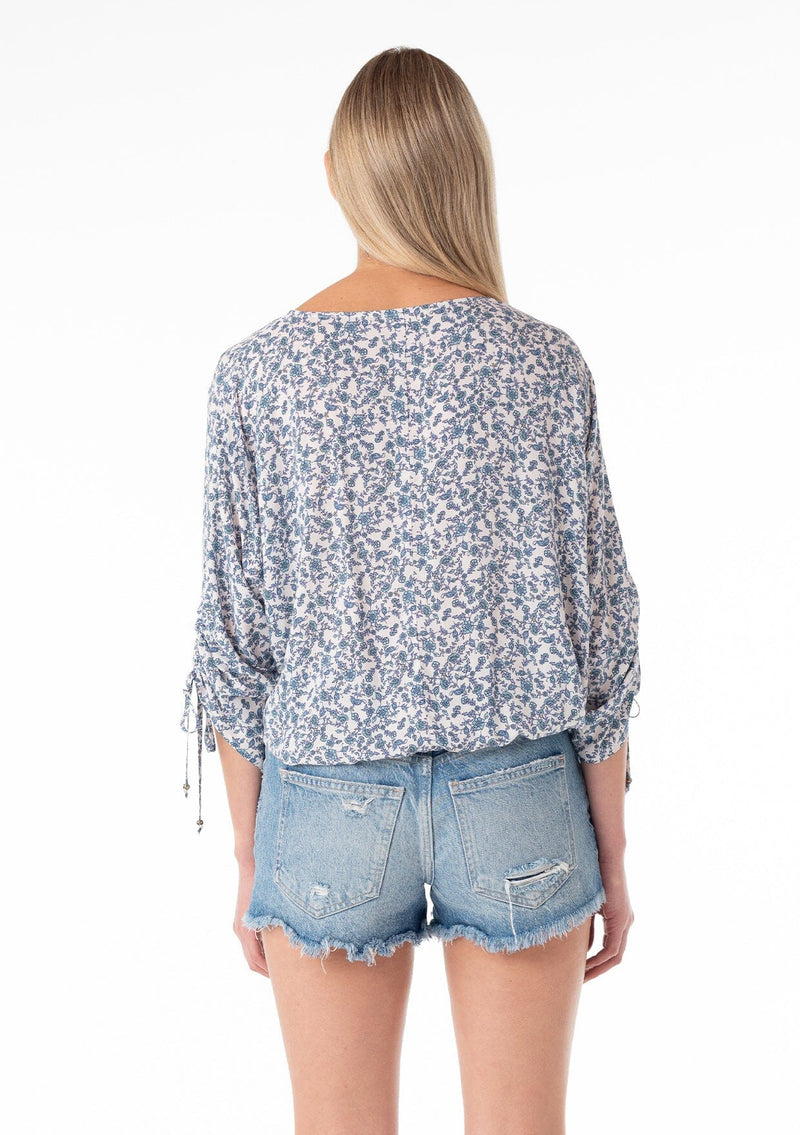 [Color: Ivory/Blue] A back facing image of a blonde model wearing a bohemian blouse in a blue floral print. With three quarter length sleeves, a gathered drawstring sleeve detail with ties, a v neckline, a self covered button front, and a relaxed fit. 