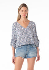 [Color: Ivory/Blue] A front facing image of a blonde model wearing a bohemian blouse in a blue floral print. With three quarter length sleeves, a gathered drawstring sleeve detail with ties, a v neckline, a self covered button front, and a relaxed fit. 