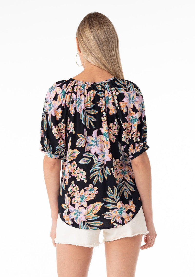 [Color: Black/Pink] A back facing image of a blonde model wearing a best selling short puff sleeve blouse in a black and pink floral print. With a self covered button front, a split v neckline, tassel ties, and a relaxed fit. 