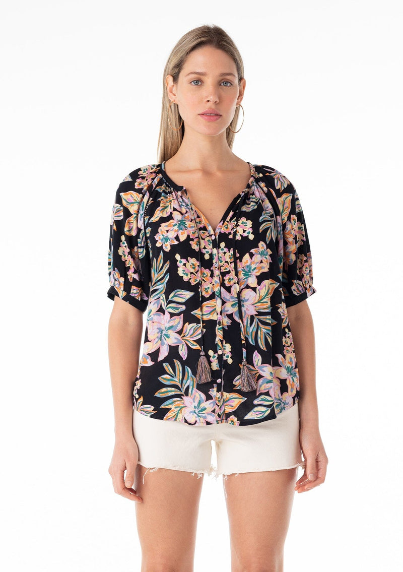 [Color: Black/Pink] A front facing image of a blonde model wearing a best selling short puff sleeve blouse in a black and pink floral print. With a self covered button front, a split v neckline, tassel ties, and a relaxed fit. 