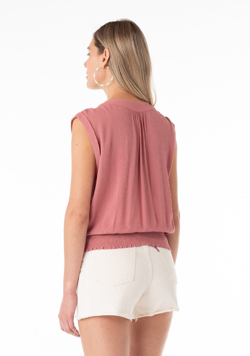 [Color: Terracotta] A three quarter back facing image of a blonde model wearing a bohemian resort top in terracotta pink. With short cap sleeves, a surplice v neckline, and a smocked elastic waist with embroidered stitch details. 