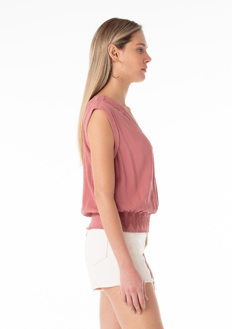 [Color: Terracotta] A side facing image of a blonde model wearing a bohemian resort top in terracotta pink. With short cap sleeves, a surplice v neckline, and a smocked elastic waist with embroidered stitch details. 