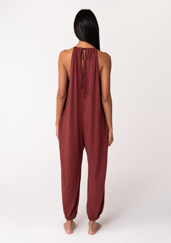 [Color: Rust] A back facing image of a brunette model wearing a cozy sleeveless, French terry jumpsuit with tie back detail, side pockets, and deep v neckline. A lounge jumpsuit in dark red. 