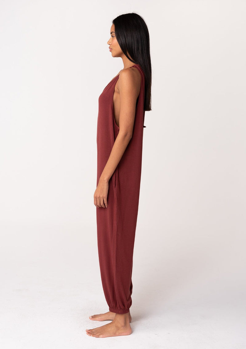 [Color: Rust] A side facing image of a brunette model wearing a cozy sleeveless, French terry jumpsuit with tie back detail, side pockets, and deep v neckline. A lounge jumpsuit in dark red. 