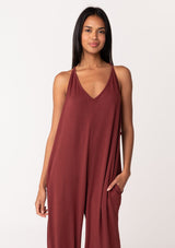 [Color: Rust] A close up front facing image of a brunette model wearing a cozy sleeveless, French terry jumpsuit with tie back detail, side pockets, and deep v neckline. A lounge jumpsuit in dark red. 