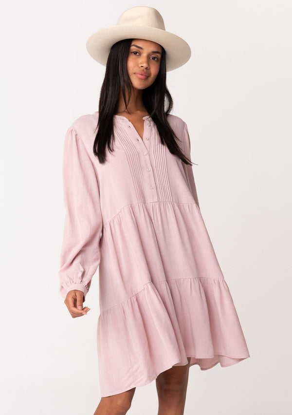 [Color: Vintage Rose] A front facing image of a brunette model wearing a linen blend pink baby doll mini dress. Features delicate pin tuck details, a button front, a flowy tiered skirt, and long voluminous sleeves with a button cuff closure.