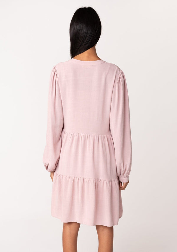 [Color: Vintage Rose] A back facing image of a brunette model wearing a linen blend pink baby doll mini dress. Features delicate pin tuck details, a button front, a flowy tiered skirt, and long voluminous sleeves with a button cuff closure.