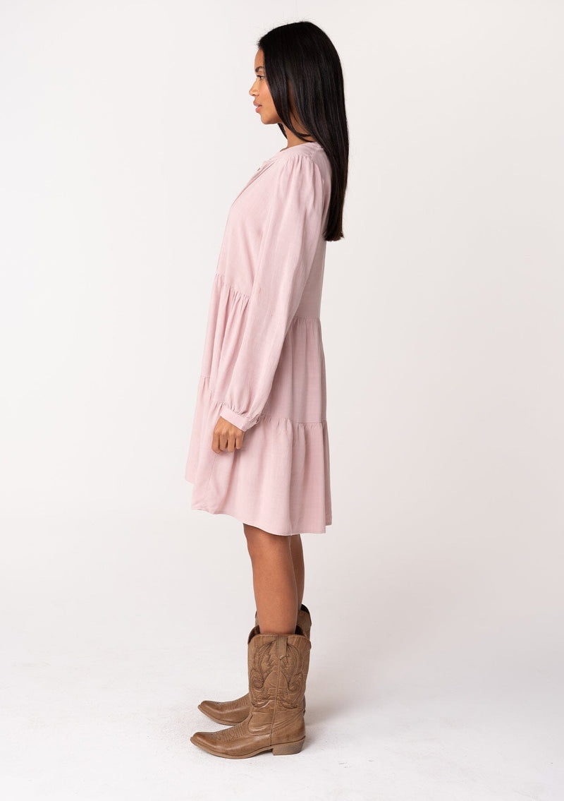 [Color: Vintage Rose] A side facing image of a brunette model wearing a linen blend pink baby doll mini dress. Features delicate pin tuck details, a button front, a flowy tiered skirt, and long voluminous sleeves with a button cuff closure.