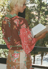 [Color: Rose/Mocha] A woman wearing a pink floral duster length robe with half length kimono sleeves, a self tie waist belt, and a ruffle trimmed tiered hemline.