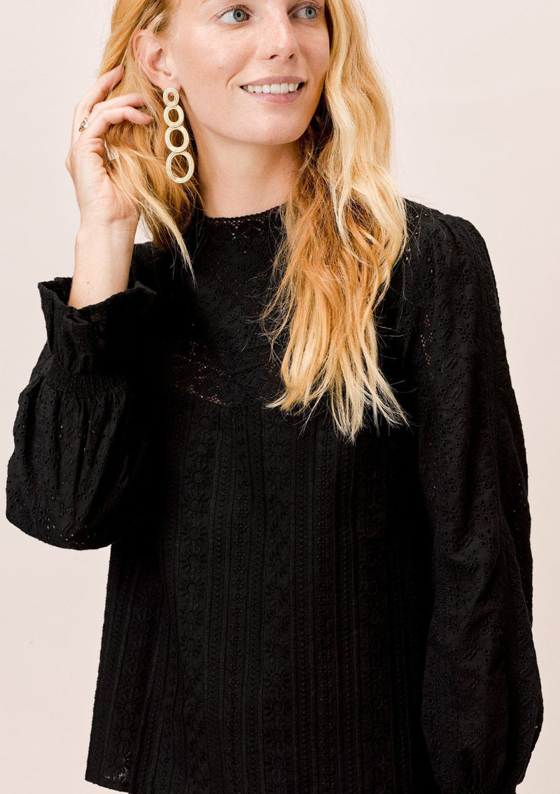 [Color: Jet Black] A blonde model wearing a black long sleeve Victorian style top with eyelet, mixed trim, mocked neckline, and smocked cuffs. 