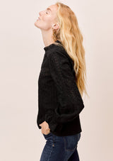 [Color: Jet Black] A blonde model wearing a black long sleeve Victorian style top with eyelet, mixed trim, mocked neckline, and smocked cuffs. 