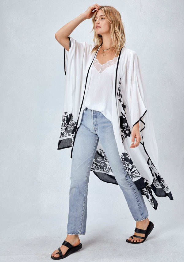 [Color: Black/White] Blonde model wearing a white bohemian kimono cover up with black border and unique black floral print. Styled over a white camisole tank top, lightwash denim jeans and black strappy sandals
