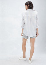 [Color: Natural/Red/Blue] A back facing image of a brunette model wearing a yarn dyed stripe shirt with subtle red stripe accents. Featuring a classic collar neckline, 3/4 length rolled sleeves, and a high low hemline. A relaxed and cool shirt for the warmer weather. 