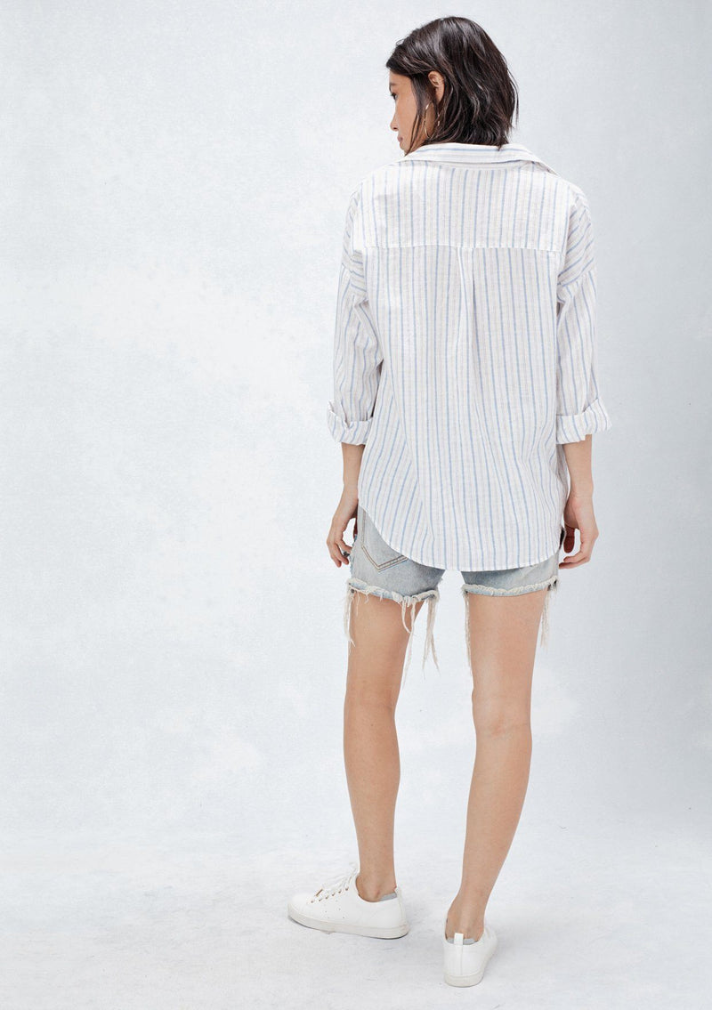 [Color: Natural/Red/Blue] A back facing image of a brunette model wearing a yarn dyed stripe shirt with subtle red stripe accents. Featuring a classic collar neckline, 3/4 length rolled sleeves, and a high low hemline. A relaxed and cool shirt for the warmer weather. 