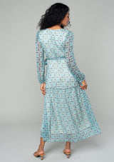 [Color: Cream/Navy] A back facing image of a brunette model wearing a flowy sheer chiffon bohemian maxi wrap dress in a navy blue and cream floral print. With long sleeves, a ruffle trimmed high low skirt, a v neckline, and a side tie waist closure. 