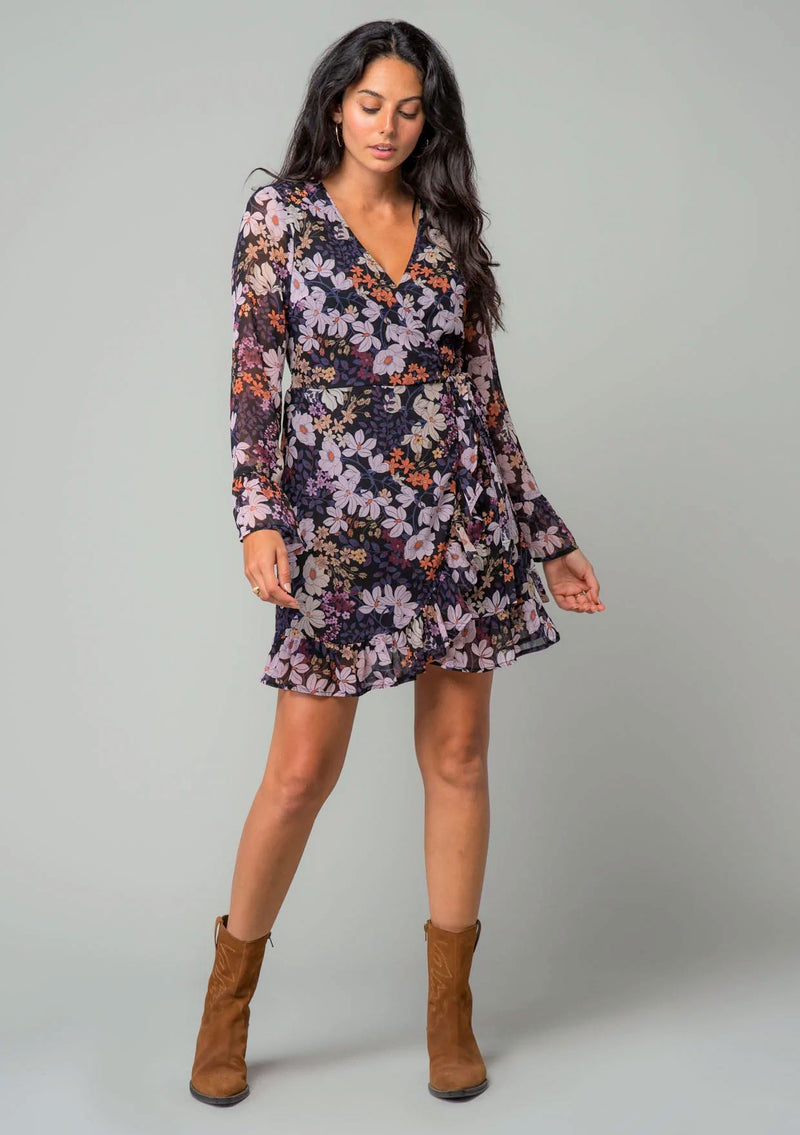 [Color: Black/Dusty Rose] A front facing image of a brunette model wearing a chiffon bohemian mini wrap dress in a black and purple floral print. With long bell sleeves, a ruffled hemline, and a side tie closure.