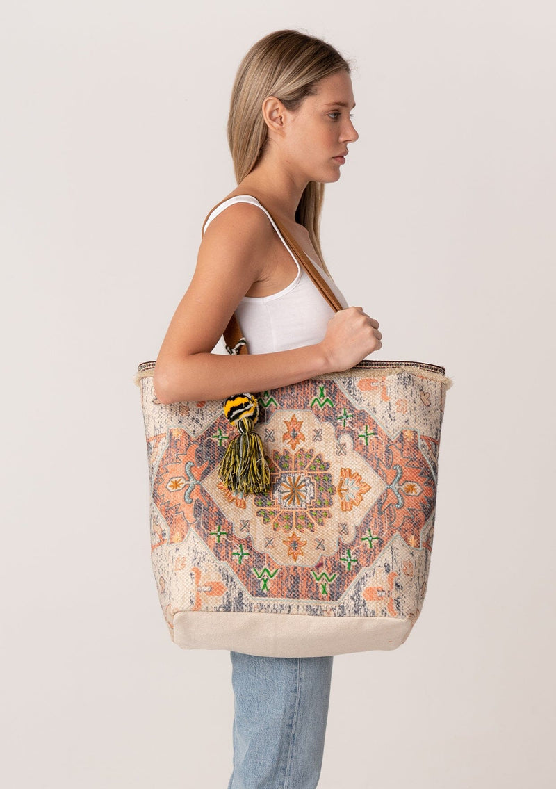 [Color: Natural/Cognac] A blonde model carrying a bohemian tote bag with tapestry design, an oversize pom tassel accent, two straps, and a magnetic closure. 