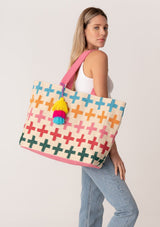 [Color: Natural/Pink] A blonde model carrying a bohemian extra large tote bag with multi color cross design throughout. With two pink straps, an extra large tassel accent, and a magnetic closure. 