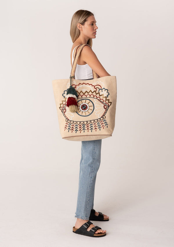 [Color: Natural/Midnight] A blonde model carrying a bohemian tote bag with a beaded eye motif. With an extra large tassel accent, two straps, and a magnetic closure. 