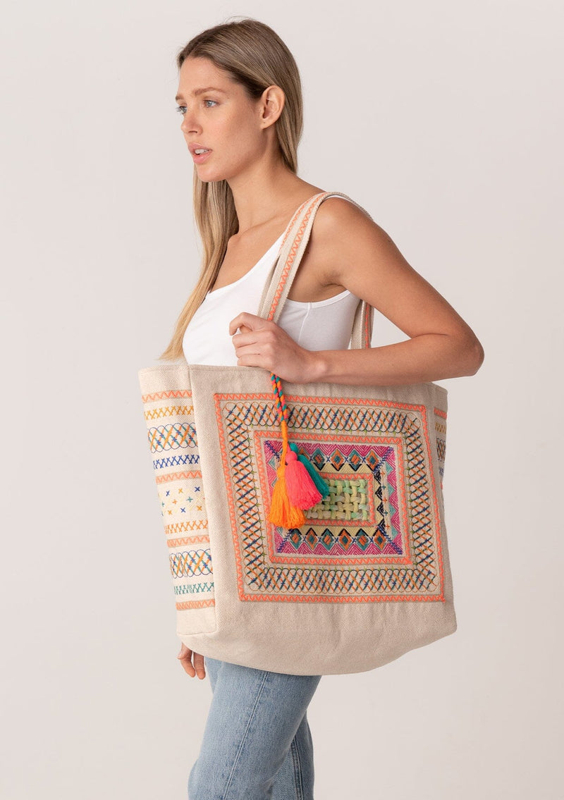 [Color: Natural/Neon Coral] A blonde model carrying a bohemian large tote bag with center lattice detail, embroidery, extra large tassel accent, and two straps. With a magnetic closure. 