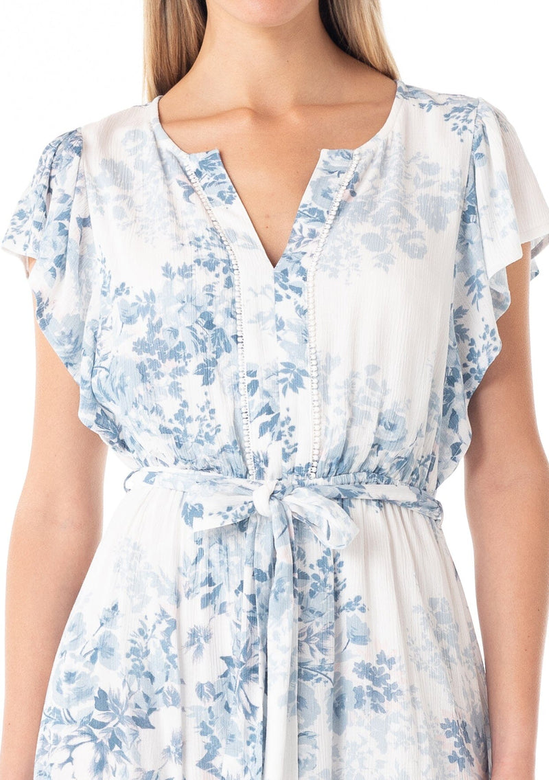 [Color: Ivory/Light Blue] A close up front facing image of a blonde model wearing a pretty spring mini dress in a white and light blue floral print. With short ruffled sleeves, a tiered mini skirt, a split v neckline, a self tie waist belt, and delicate mini pom trim throughout. 