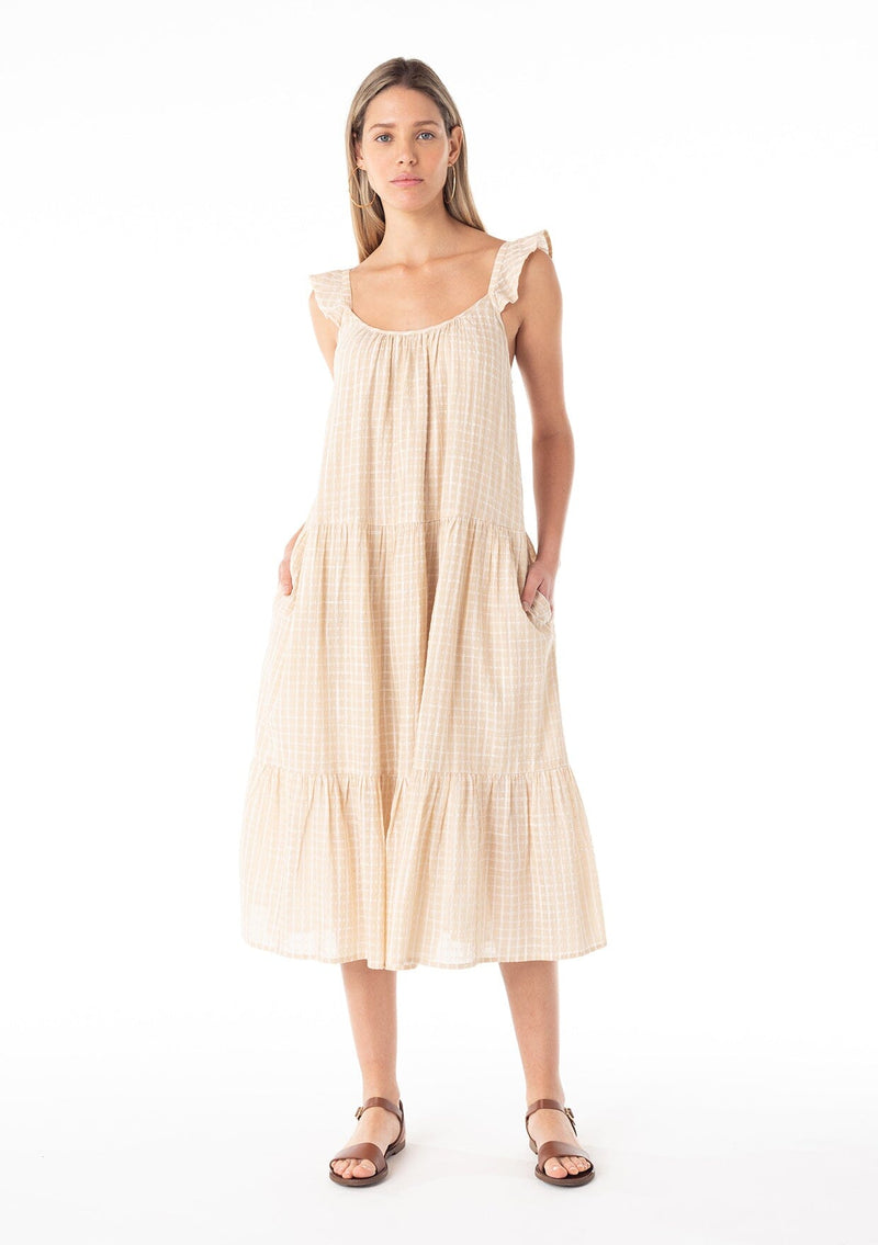 [Color: Natural] A front facing image of a blonde model wearing a casual bohemian mid length dress in a natural cotton seersucker check print. With adjustable ruffle straps, a scoop neckline in the front and back, side pockets, and a tiered flowy silhouette.