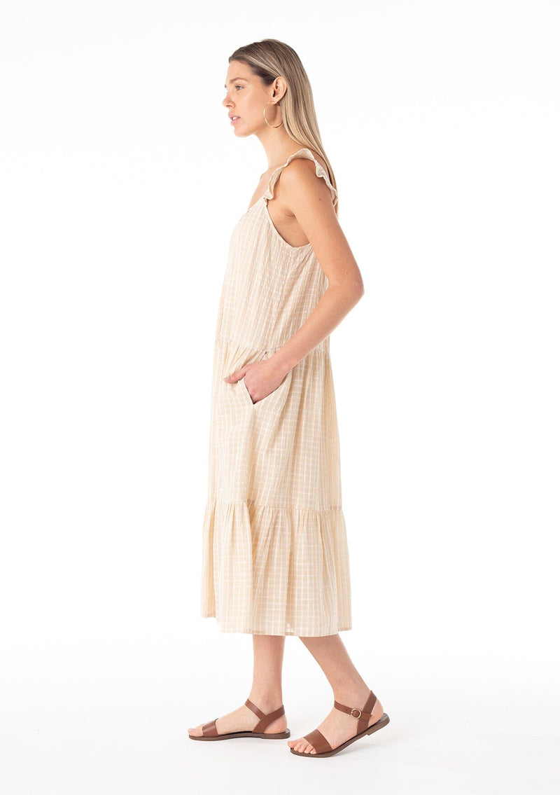 [Color: Natural] A side facing image of a blonde model wearing a casual bohemian mid length dress in a natural cotton seersucker check print. With adjustable ruffle straps, a scoop neckline in the front and back, side pockets, and a tiered flowy silhouette.
