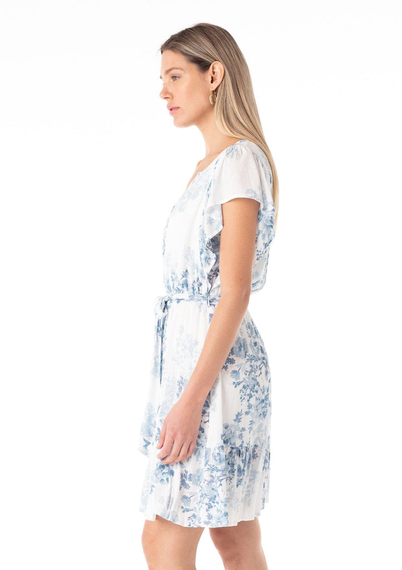 [Color: Ivory/Light Blue] A side facing image of a blonde model wearing a pretty spring mini dress in a white and light blue floral print. With short ruffled sleeves, a tiered mini skirt, a split v neckline, a self tie waist belt, and delicate mini pom trim throughout. 
