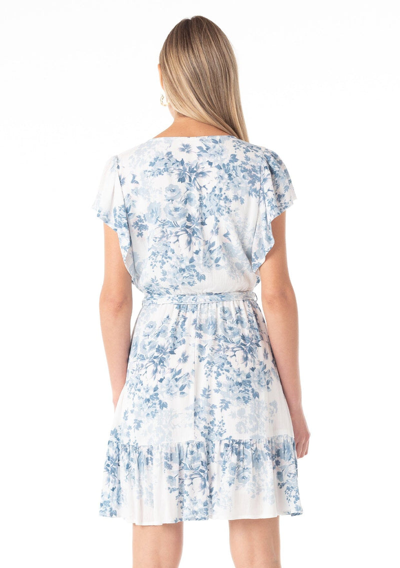 [Color: Ivory/Light Blue] A back facing image of a blonde model wearing a pretty spring mini dress in a white and light blue floral print. With short ruffled sleeves, a tiered mini skirt, a split v neckline, a self tie waist belt, and delicate mini pom trim throughout. 